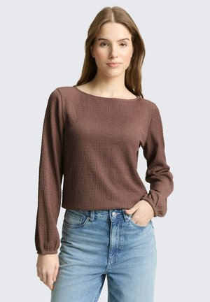 Młoda kobieta w brązowej, teksturowanej bluzce z długim rękawem i jasnobłękitnych, wysokich dżinsach, stojąca z jedną ręką w kieszeni na jednolitym tle.