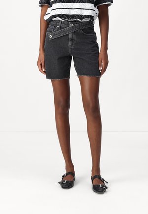 Person iført sort denimshorts med høj talje og asymmetrisk bælte, en sort-hvid mønstret top og sorte sko med spænde.
