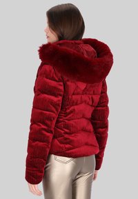 Červená péřová bunda s kapucí s umělým chlupem, žebrovanou texturou a prošívaným designem. Nošena se lesklými kovově zlatými kalhotami. Bez viditelných zapínání.