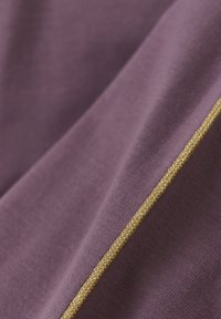 Fioletowy materiał o gładkiej strukturze, z złotym plecionym wykończeniem wzdłuż jednej krawędzi, podkreślający czysty, minimalistyczny design.