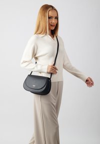 Black leather crossbody bag with a rounded flap, golden hardware detail, and an adjustable strap, paired with a beige sweater and light pants.