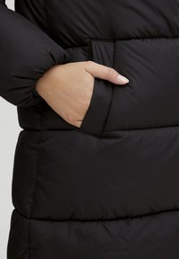 Svart pufferjakke med quiltet tekstur. Har en sidelomme og tettsittende ermer. Glatt, blank overflate.