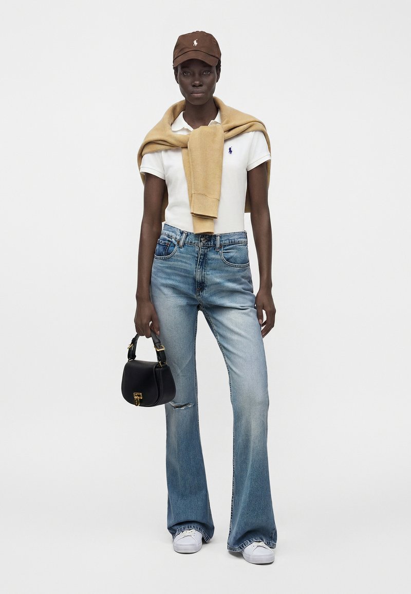 Femme portant un polo blanc, un pull beige drapé sur les épaules, un jean évasé bleu, des baskets blanches, une casquette marron, portant un petit sac à main noir.