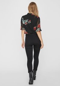 Blusa nera a fiori con maniche corte, ornata da motivi verdi e rossi, abbinata a jeans skinny neri e stivaletti con un design testurizzato.