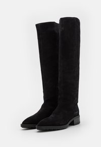 Paire de bottes noires en daim montant jusqu'au genou, avec des talons blocs bas et des bouts arrondis, posées debout sur un fond blanc.