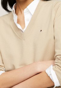 Beżowy sweter w serek z dzianiny, z małym haftowanym logo na piersi. Nałożony na białą koszulę z kołnierzykiem.