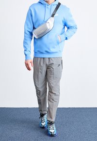 Modrá mikina s kapucí, šedé kalhoty a vícebarevné boty s výrazným vzorem. Stříbrná ledvinka nošená přes hruď.