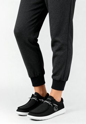 Schwarze Slipper mit weißen Sohlen und grauen elastischen Schnürsenkeln, getragen mit dunkelgrauen, umgeschlagenen Jogginghosen vor weißem Hintergrund.