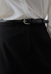 Čierne nohavice na mieru s hladkou textúrou, sú vybavené tenkým čiernym opaskom a striebornou kovovou prackou, v kombinácii s bielou košeľou na gombíky.