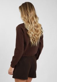 Maglione marrone texturizzato con maniche lunghe e polsini a coste, abbinato a pantaloncini scuri. La vista mostra il retro dell'outfit e capelli lunghi e ondulati.