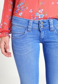 Modré džínové kalhoty s přiléhavým střihem, se dvěma kovovými knoflíky v pase a předními kapsami s jemnými švy.