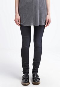 Svarta tajta jeans med en slät textur, kombinerat med en grå oversized topp och svarta polerade skor med dubbla spännen.