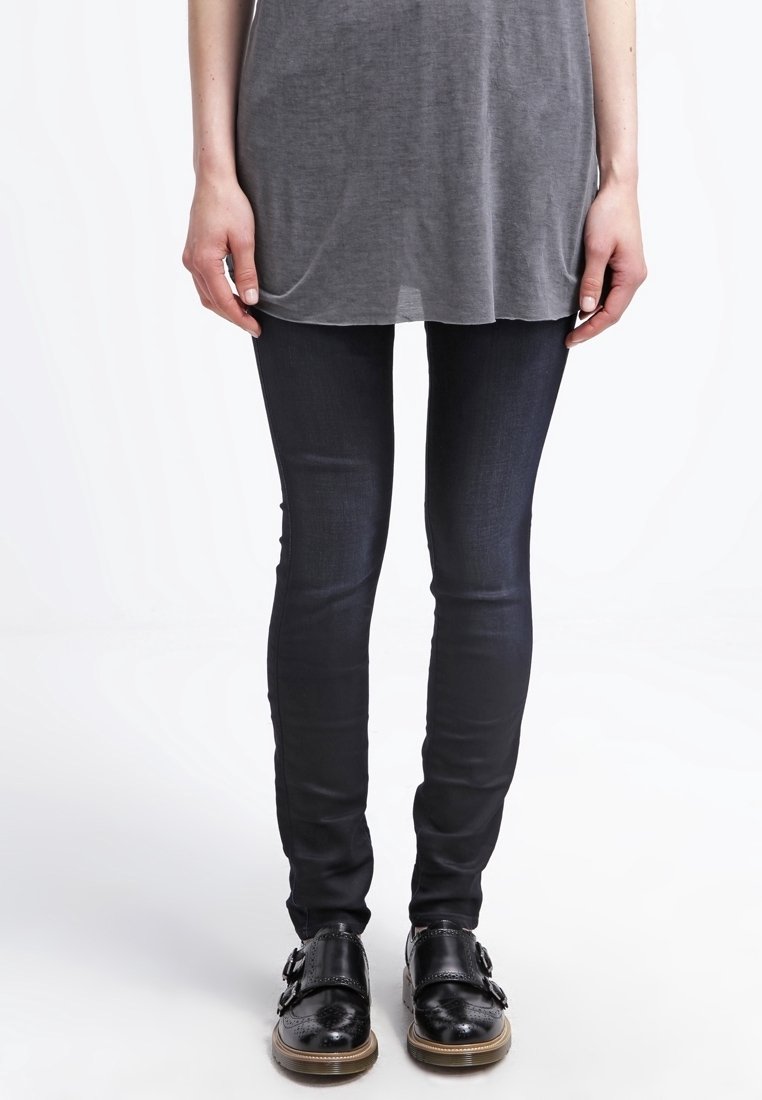 Svarta tajta jeans med en slät textur, kombinerat med en grå oversized topp och svarta polerade skor med dubbla spännen.