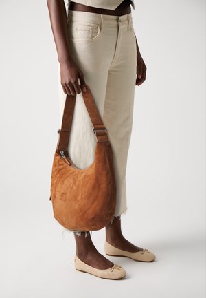 Bruine suède schoudertas met een gebogen vorm, metalen accenten en een brede band. Gecombineerd met off-white wijde broek en beige flats.