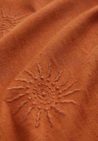 Terrakotta-Stoff mit weicher Textur, der aufwendige Sonnenstickerei in einem kreisförmigen Design mit ausstrahlenden Linien und Punkten aufweist.