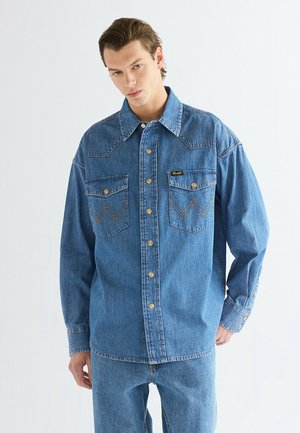 Junger Mann, der ein blaues Jeanshemd mit geknöpften Brusttaschen und passende blaue Jeans trägt, steht vor einem schlichten hellen Hintergrund.