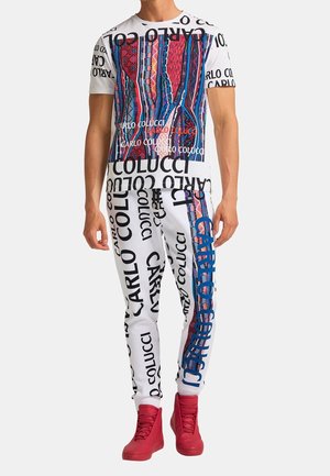 Valkoinen lyhythihainen t-paita, jossa on värikäs kuvio etuosassa, yhdistettynä valkoisiin housuihin, joissa on rohkeaa mustaa tekstiä ja sinisiä yksityiskohtia. Punaiset korkeakorkoiset kengät.