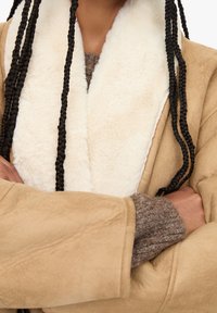 Beige faux-velour frakke med en fluffy hvid krave, store ærmer og en tekstureret brun striktrøje under.