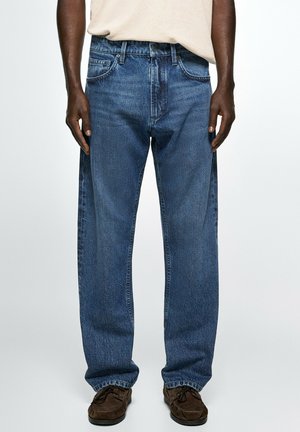 Mann trägt locker sitzende blaue Jeans, beiges Hemd und braune Schuhe, steht mit locker hängenden Armen an den Seiten vor einem schlichten Hintergrund.