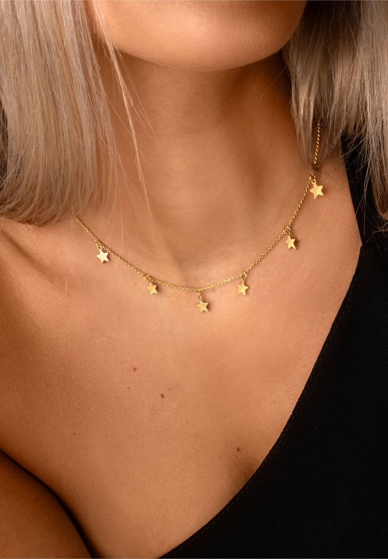Collier chaîne en or avec cinq petits pendentifs en forme d’étoile porté autour du cou et sur l’épaule d’une femme, partiellement recouvert par un vêtement noir.