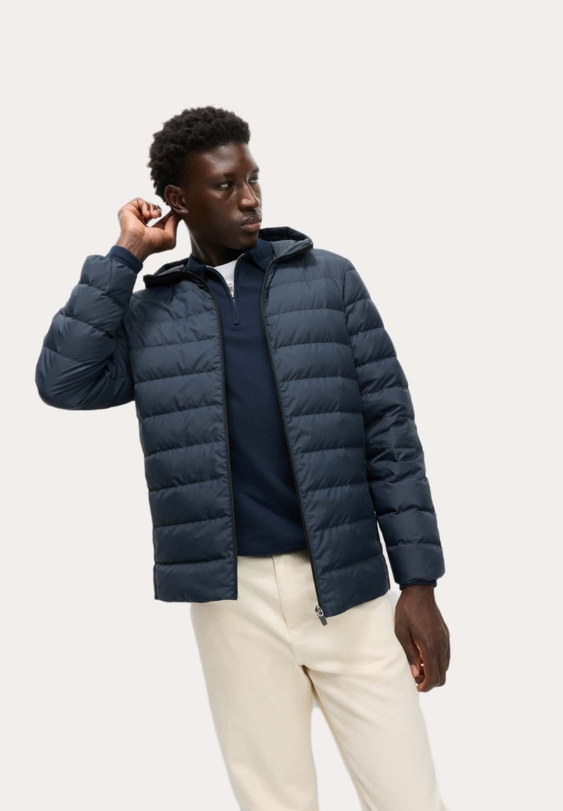 Giubbotto imbottito di un blu navy scuro, con sezioni trapuntate, cappuccio e chiusura con zip anteriore, abbinato a un top navy e pantaloni chiari.