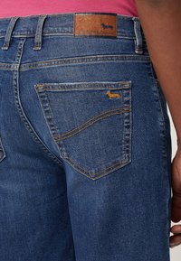 Donkerblauwe denim jeans met oranje stiksels, voorzien van een achterzakontwerp met een klein geborduurd teckel-logo en een leren label op de taille.