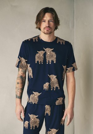 Man with shoulder-length hair wearing navy blue outfit with matching tops and bottoms featuring beige shaggy highland cow patterns.