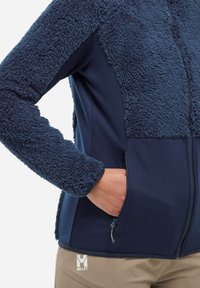 Osoba w granatowej polarowej kurtce z ręką w zapinanej kieszeni, w zestawieniu z beżowymi spodniami z białym logo.