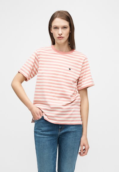 Γυναίκα που φοράει ροζ και λευκή ριγέ μπλούζα και μπλε τζιν, στέκεται με το ένα χέρι στην τσέπη της μπροστά σε απλό λευκό φόντο.