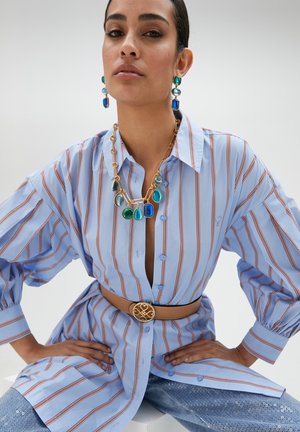 Camisa de rayas azul claro con mangas amplias, combinada con un cinturón beige que presenta un acento dorado. Joyería audaz en tonos azules y verdes.