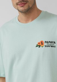 Jasno miętowy t-shirt z bawełny z nadrukowanym grafiką papai oraz napisem "PAPAJOWA PASTA DO SURFINGU" w kolorze czarnym i pomarańczowym.