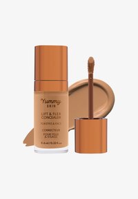 Flytende concealer i en gjennomsiktig glassflaske med en bronsefarget hette. Har en myk applikator med en kremet brun produktprøve ved siden av.