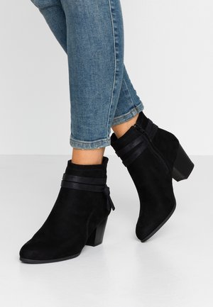 Jambes en jean bleu portant des bottines en daim noires avec talons carrés et fermetures éclair latérales, debout sur un sol blanc.