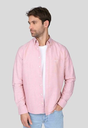 Chemise à manches longues rose clair avec des rayures verticales blanches, col boutonné et poche poitrine. Portée par-dessus un t-shirt blanc, coupe décontractée.