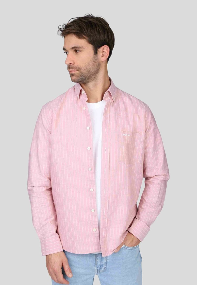 Chemise à manches longues rose clair avec des rayures verticales blanches, col boutonné et poche poitrine. Portée par-dessus un t-shirt blanc, coupe décontractée.