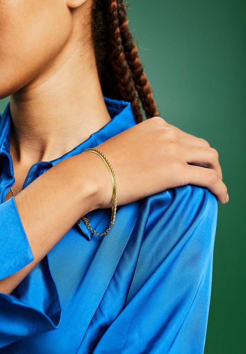 Gouden armband met een gedraaide design gedragen om een blote pols, gecombineerd met een levendige blauwe satijnen shirt. Groene achtergrond versterkt de compositie.