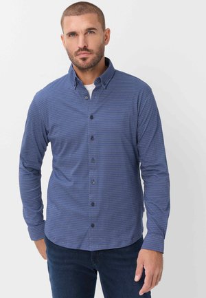Mand med kort hår og skæg iført en knappet blå mønstret skjorte over en hvid undertrøje og mørkeblå jeans, stående mod en ensfarvet baggrund.