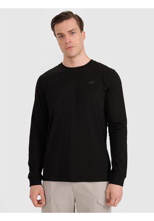 Hombre con camisa negra de manga larga y pantalones beige claro de pie frente a un fondo liso gris claro.