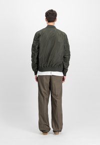 Jachetă bomber de culoare verde măslinie, cu manșete cu nervuri, dintr-un material ușor și cu umerii căptușiți. Pur tată peste o cămașă gri deschis și pantaloni căprui.