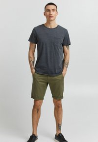 T-shirt gris à manches courtes avec détail de poche, associé à un short vert olive. Des baskets noires complètent l'ensemble. Fond uni.