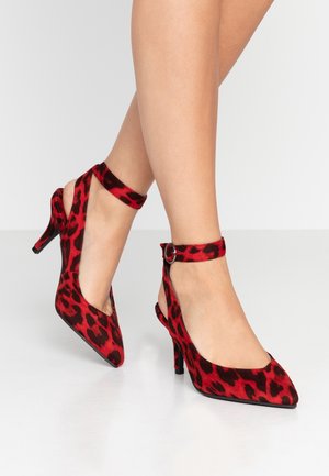 Frau trägt rot-schwarze High Heels mit Leopardenmuster und Knöchelriemen und steht auf einem weißen Boden. Es sind nur die Beine und Schuhe sichtbar.