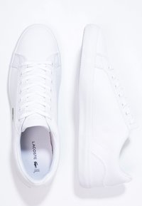 Paire de baskets basses Lacoste blanches, l'une vue de dessus avec les lacets, l'autre vue de côté, sur fond blanc.