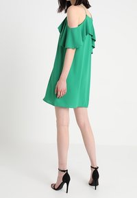 Abito verde a spalle scoperte con volant sovrapposti, tessuto leggero e vestibilità ampia, abbinato a sandali neri con tacco alto.