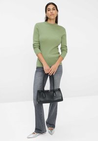 Un pull vert côtelé, un jean gris évasé et un sac à main en cuir noir. Les chaussures sont argentées avec un bout pointu. Fond neutre.