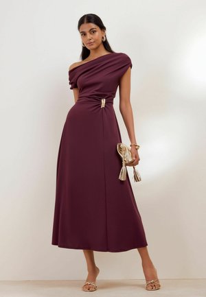 Frau trägt ein burgunderrotes Midi-Kleid mit einem Schulter, hält eine goldene Clutch und trägt goldene Sandalen, Ohrringe und Armband.