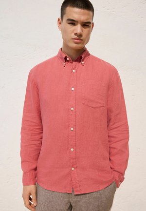 Junger Mann mit kurzen Haaren, der ein langärmeliges pinkfarbenes Hemd mit Knöpfen und grau karierte Hose trägt, vor einer schlichten hellen Wand.