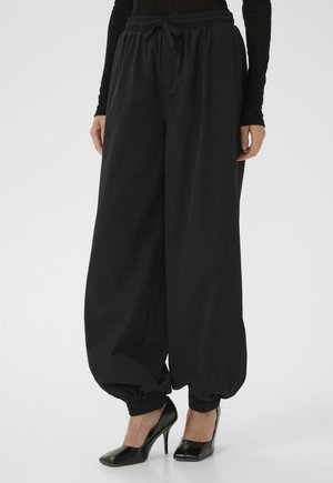 Kobieta ubrana w luźne czarne spodnie z elastycznymi mankietami i czarne buty na wysokim obcasie, stojąca na jednolitym tle.