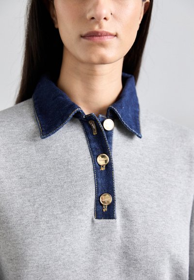 Felpa grigia con colletto in denim, caratterizzata da una chiusura con bottoni con accenti dorati e cuciture a contrasto. Texture complessivamente liscia.