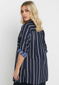 Blusa a righe blu navy con strisce verticali bianche, maniche arrotolate con un accento blu e vestibilità morbida con fessure laterali.