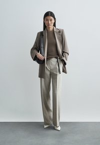 Bruine blazer over een bruine top, lichtgrijze broek met zwarte strikriem en witte schoenen met puntige neus. Kleine ronde zwarte tas met bandje.